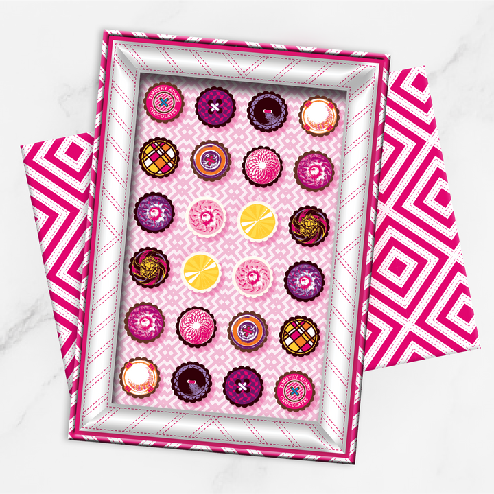 A gift box of 24 truffles with a variety of colors and patterns on the tops, presented on a pink and white checkered surface.
