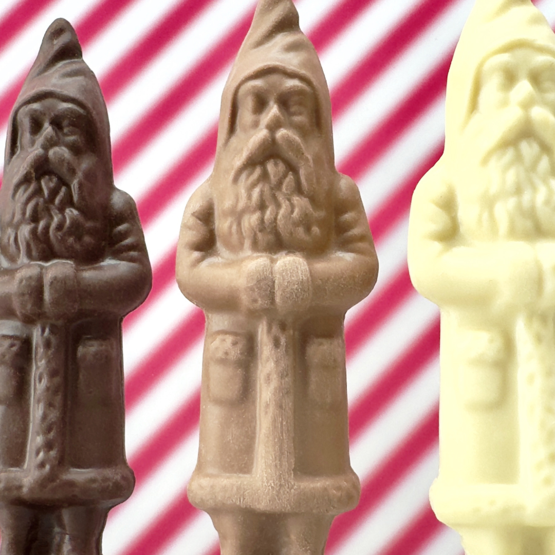 Three chocolate Santa figures in Valrhona solid dark, milk, and white chocolate standing on marble with a striped holiday backdrop.