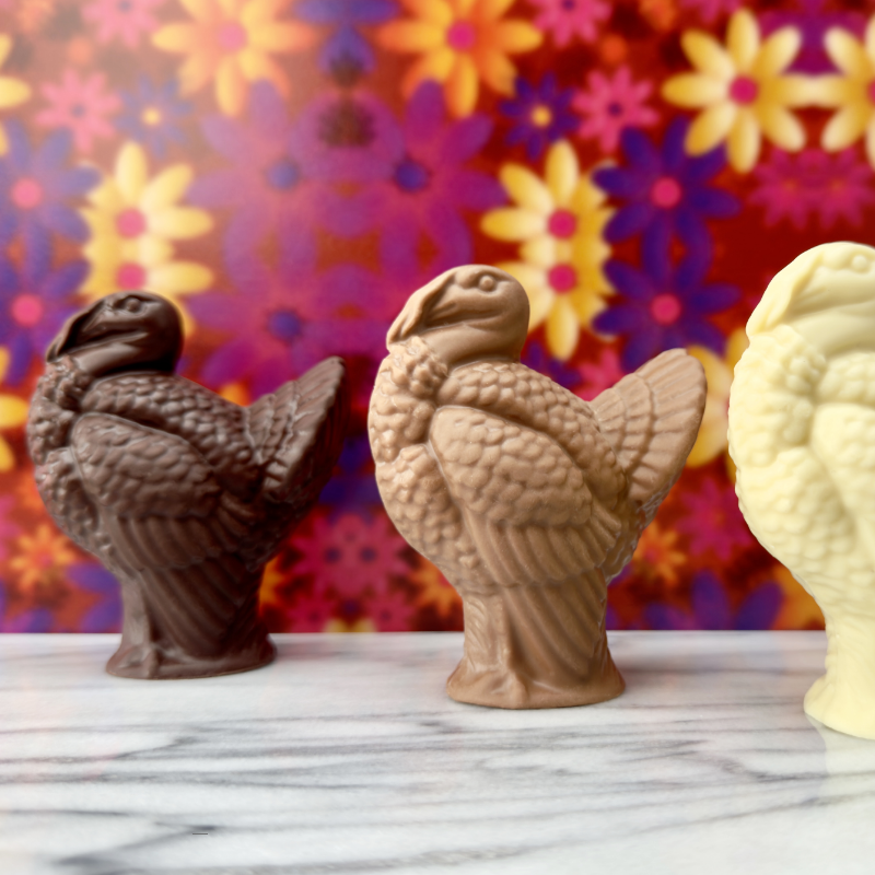 Three handcrafted chocolate turkeys—dark, milk, and white chocolate—displayed on marble with a vibrant floral backdrop, created at Timothy Adams Chocolates.