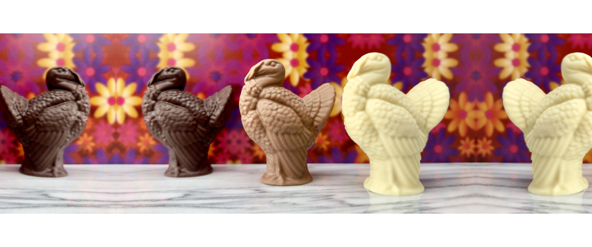 Three handcrafted chocolate turkeys—dark, milk, and white chocolate—displayed on marble with a vibrant floral backdrop, created at Timothy Adams Chocolates.