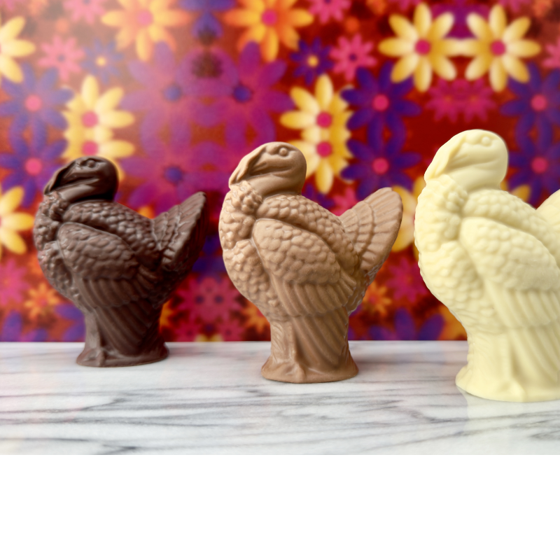 Three handcrafted chocolate turkeys—dark, milk, and white chocolate—displayed on marble with a vibrant floral backdrop, created at Timothy Adams Chocolates.”