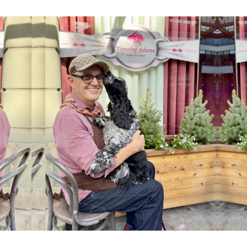 Tim the owner sits outside the shop, smiling as the black-and-white dog — Rosie, climbs into their lap to nuzzle them affectionately. Behind them is the storefront with a sign for the chocolate shop and a planter of small evergreens and flowers.